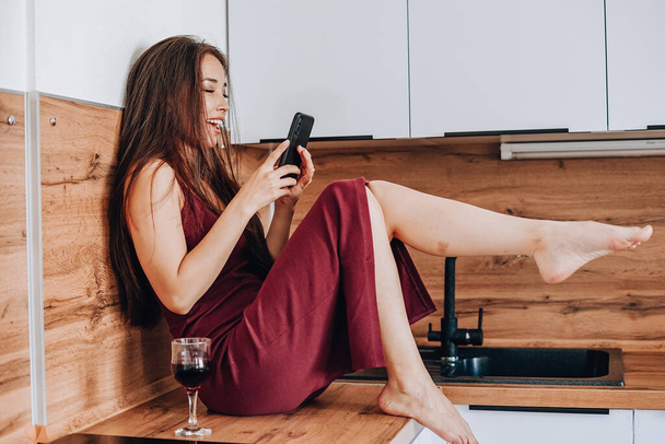 attractive Asian woman drinking wine and using a mobile phone on the countertop in the kitchen at home - Fotografie, Obrázek