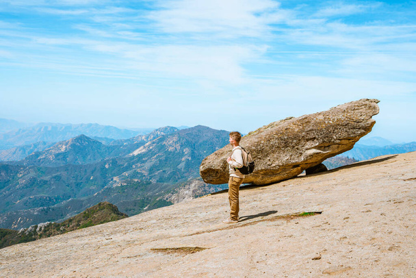 Ein junger Mann geht im Sequoia Nationalpark in den USA in den Bergen neben einem hängenden Stein wandern. Erstaunliche Landschaft von der Klippe bis zum blauen Himmel und den Bergen - Foto, Bild