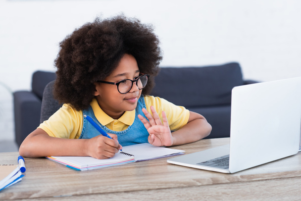 Afrikaans amerikaans meisje zwaaien hand op laptop en schrijven op notebook thuis  - Foto, afbeelding