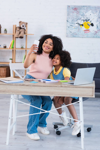 Sorridente madre e bambino afroamericano che si scattano selfie vicino a laptop e cancelleria a casa  - Foto, immagini