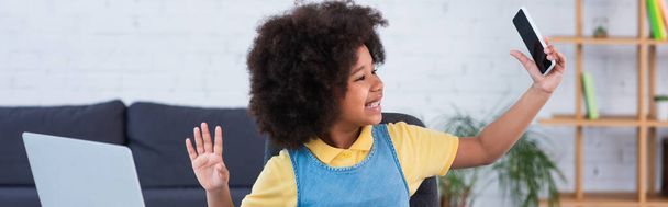 Glimlachend Afrikaans Amerikaans kind zwaait hand op laptop en houdt smartphone thuis, banner  - Foto, afbeelding