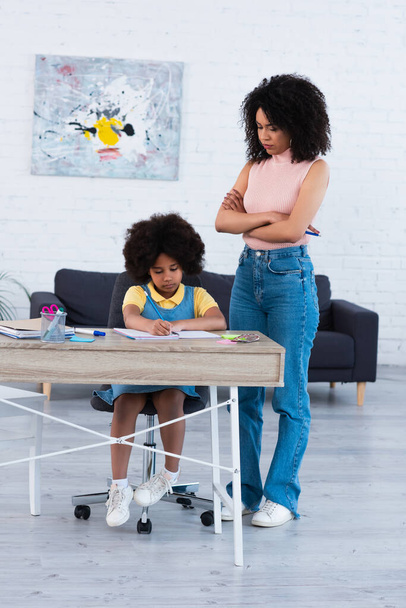 Serious african american woman looking at daughter writing on notebook at home  - Foto, Imagen