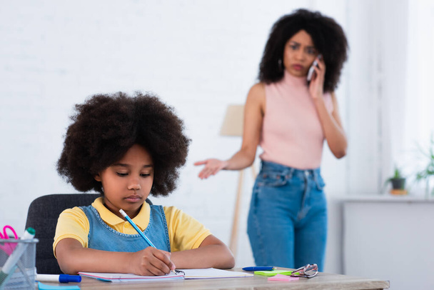 Afrykańskie amerykańskie dziecko piszące na notatniku w pobliżu rozmytej matki mówiące na smartfonie w domu  - Zdjęcie, obraz
