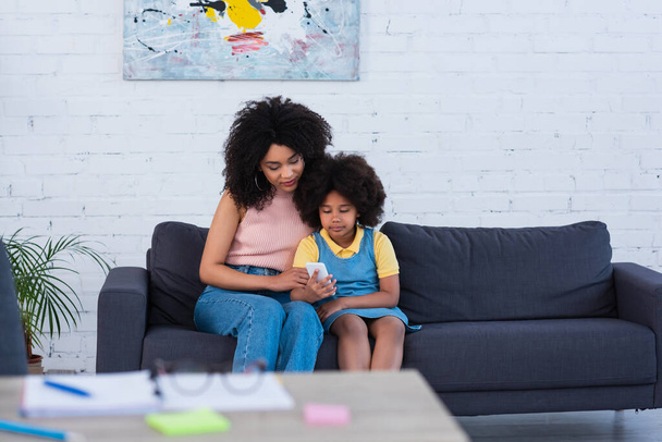 African american mother and girl using smartphone near blurred stationery at home  - 写真・画像