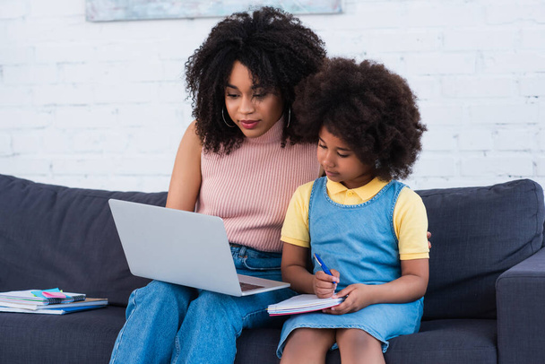 Afrika kökenli Amerikalı anne dizüstü bilgisayar tutarken, kız da online ders sırasında deftere yazı yazıyor.  - Fotoğraf, Görsel