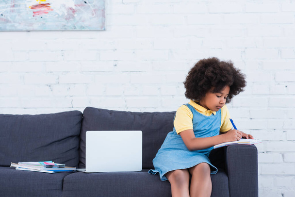 African american kid writing on notebook near laptop on couch  - Foto, Bild
