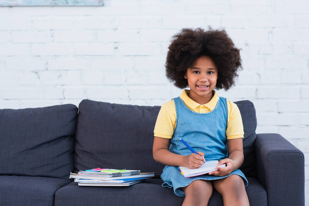African american girl smiling at camera while doing schoolwork on couch  - Фото, зображення