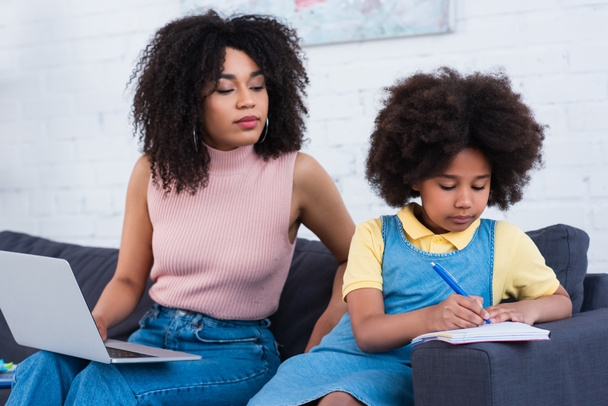 African american kid writing on notebook near blurred mother with laptop on couch  - Photo, Image