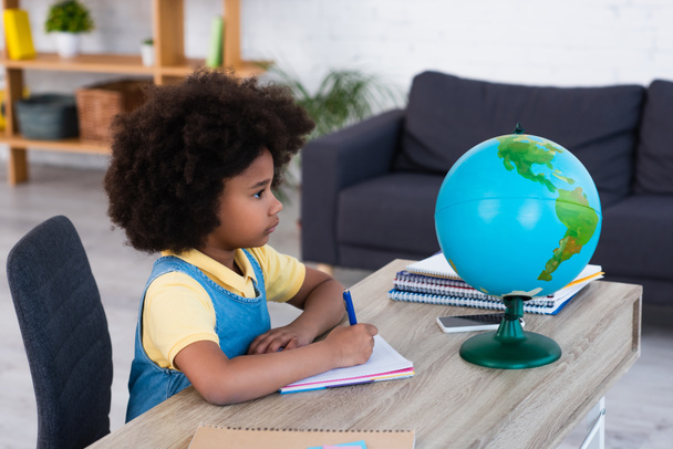 Side view of african american girl writing on notebook and looking at globe at home  - Photo, Image