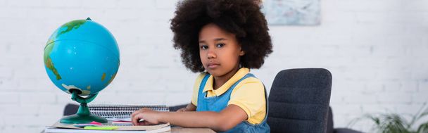 Chica afroamericana mirando a la cámara cerca de papelería y globo en la mesa, pancarta  - Foto, Imagen