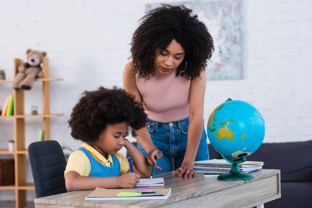 African american woman pointing at notebook while daughter writing near globe  - Foto, immagini