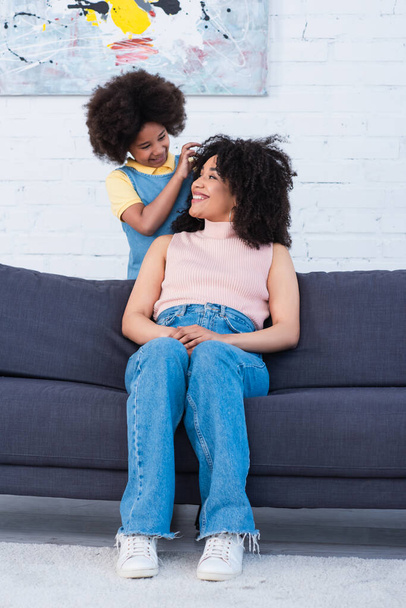 African american girl touching hair of mother on couch in living room  - Foto, Imagem