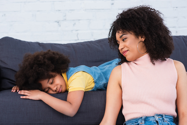 Afroamerykanka matka patrząc na śpiącej córki na kanapie w salonie  - Zdjęcie, obraz