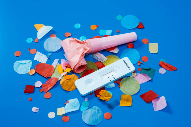 a covid-19 antigen diagnostic test device, a pink party horn and some multicolored confetti on a blue background, as a safe party set - Photo, Image
