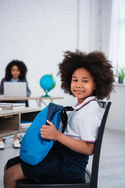 Sonriente niño afroamericano sosteniendo mochila en el aula  - Foto, Imagen