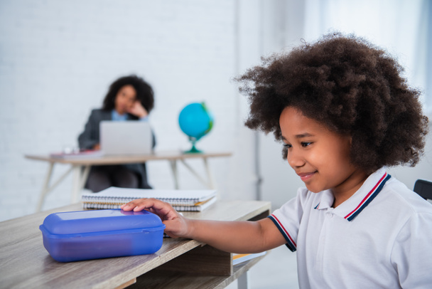Mosolygó afro-amerikai gyerek kezében ebéd doboz közelében elmosódott notebook az osztályteremben  - Fotó, kép