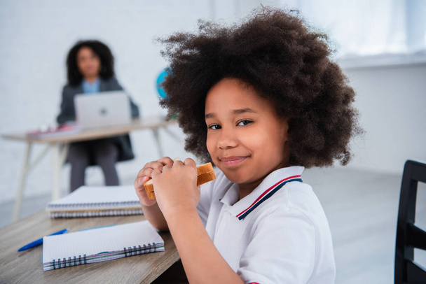 Alumno afroamericano sosteniendo sándwich cerca de la libreta en el escritorio  - Foto, Imagen