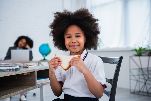 Lächelnde afrikanisch-amerikanische Schülerin hält Sandwich im Klassenzimmer  - Foto, Bild