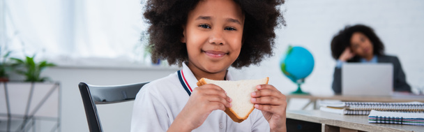 African american schoolchild holding sandwich during break in classroom, banner  - Photo, Image