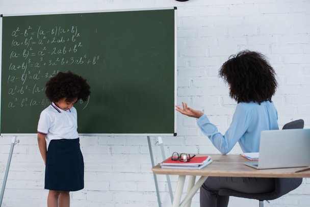 Afro-Amerikaanse leraar zitten in de buurt boos leerling naast schoolbord in de klas  - Foto, afbeelding