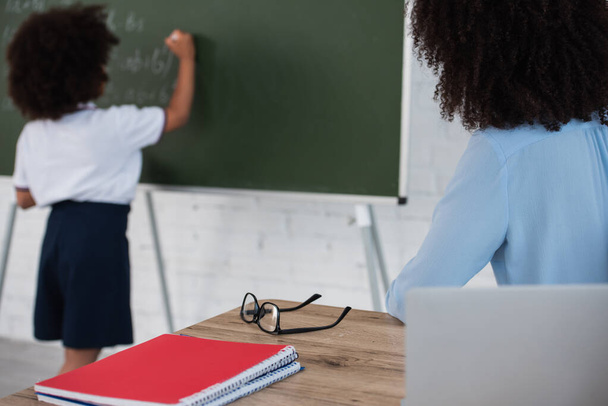 African american teacher sitting near laptop and blurred pupil writing on chalkboard  - Photo, image
