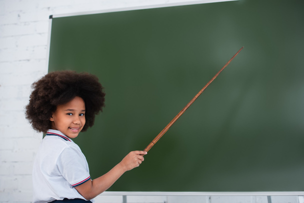 Mosolygó afro-amerikai iskolás gyerek néz kamera és rámutatva kréta az osztályteremben  - Fotó, kép