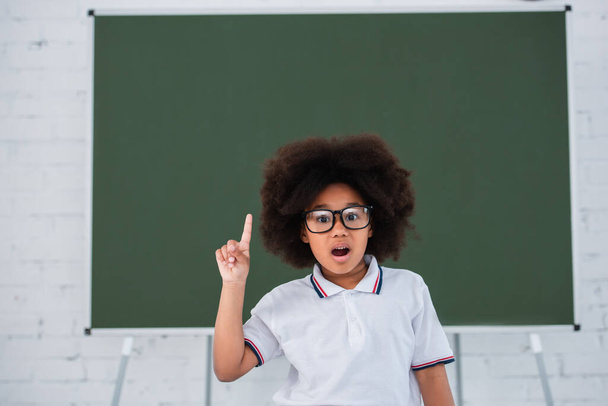 Astonished african american pupil having idea near blurred chalkboard in classroom  - Valokuva, kuva