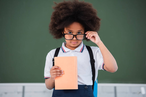 African american pupil holding eyeglasses and book near chalkboard in classroom  - Fotoğraf, Görsel