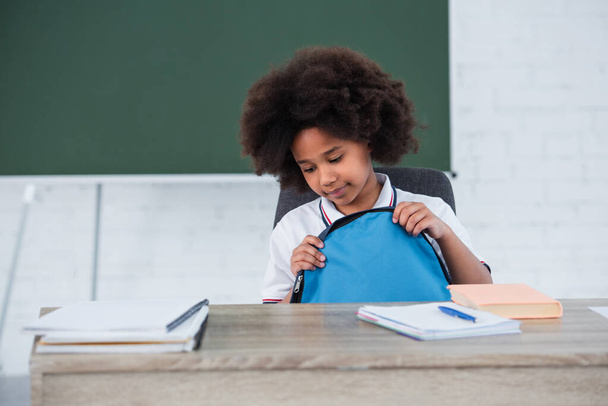 African american kid holding backpack near notebooks on desk in classroom  - Фото, изображение
