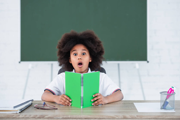 Niño afroamericano asombrado sosteniendo cuaderno en el aula  - Foto, imagen