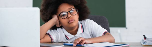 Uitgeput Afrikaans Amerikaans schoolkind in bril kijkend naar camera in de buurt van laptop en briefpapier, banner  - Foto, afbeelding