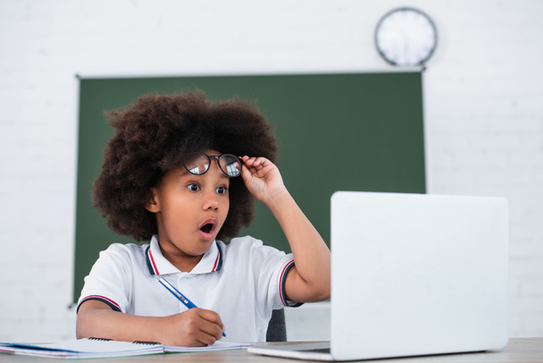 Bambino africano americano stupito che tiene gli occhiali vicino a laptop e cancelleria sfocati  - Foto, immagini