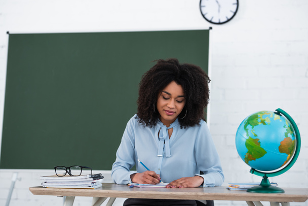African american teacher writing on notebook near globe in classroom  - Foto, immagini