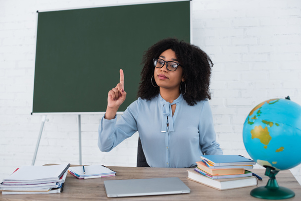 African american teacher having idea near laptop, blurred globe and stationery in classroom  - Photo, Image