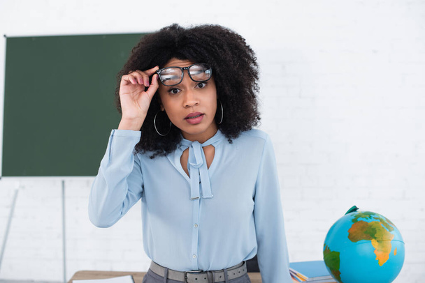 Amazed african american teacher holding eyeglasses and looking at camera in classroom  - Photo, Image