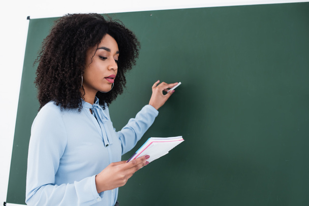 African american teacher with notebook writing on chalkboard in classroom  - 写真・画像