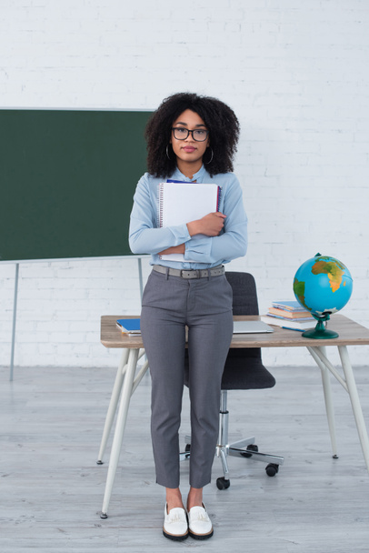 African american teacher in eyeglasses holding notebooks near laptop and globe in school  - Foto, imagen