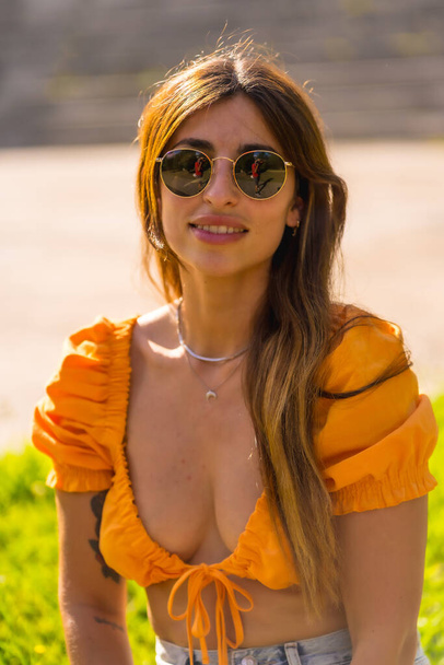 Lifestyle of a young caucasian brunette enjoying the summer in a park in the city. Girl with sunglasses and an orange t-shirt - Фото, изображение
