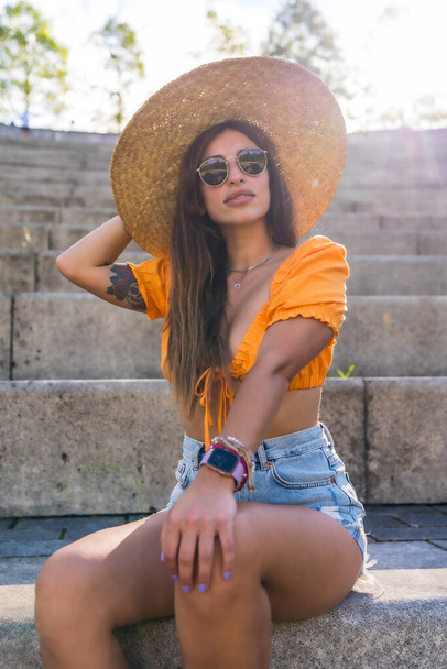 Lifestyle of a young caucasian brunette walking in the summer in a park in the city. Portrait of the girl with a hat at sunset, vertical photo - Zdjęcie, obraz