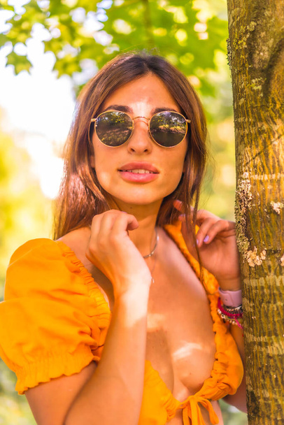 Lifestyle of a young caucasian young brunette in a park in summer next to a tree in nature. Girl with glasses smiling, looking at camera - 写真・画像