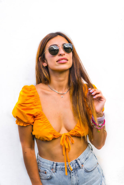 Lifestyle of a young caucasian brunette with orange t-shirt, sunglasses and short pants on a white background, with copy space and sticks. With a sweet and seductive look - Zdjęcie, obraz