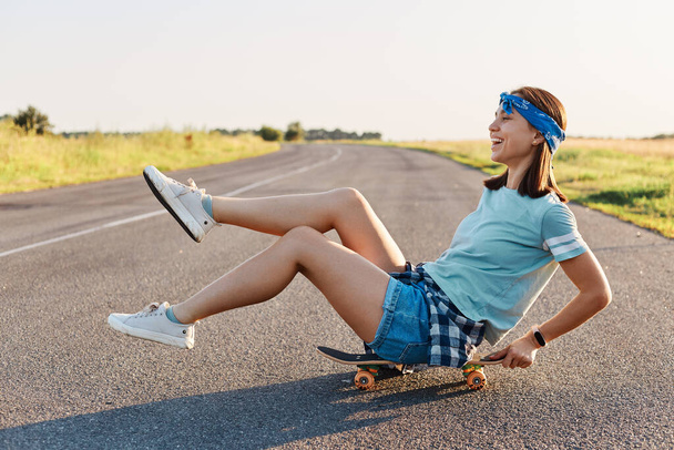 Side view of happy emotional woman sitting on skateboard on the street with raised legs, overjoyed female wearing casual clothing having fun outdoors, positive lifestyle. - Φωτογραφία, εικόνα