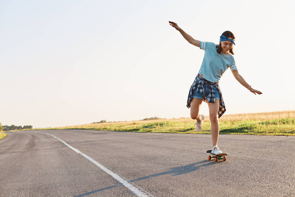 Full length portrait of happy young adult female skateboarding outdoor on asphalt road alone, raised arms, wearing casual style clothing, expressing positive emotions. - Foto, afbeelding