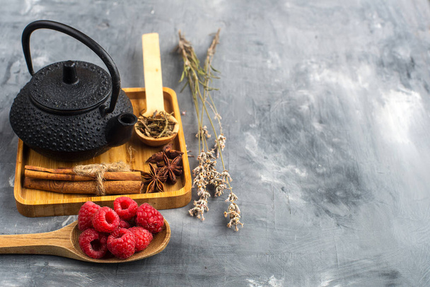 cast iron teapot with tea on a wooden tray and a spoon of raspberries on a gray background. space for text - Foto, Imagen