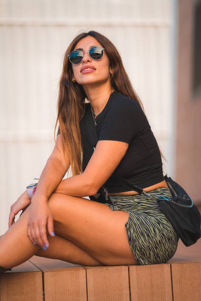 Street Style of an attractive young caucasian brunette sitting in the city with sunglasses, enjoying a veranao with a white wall background, vertical photo - Фото, зображення