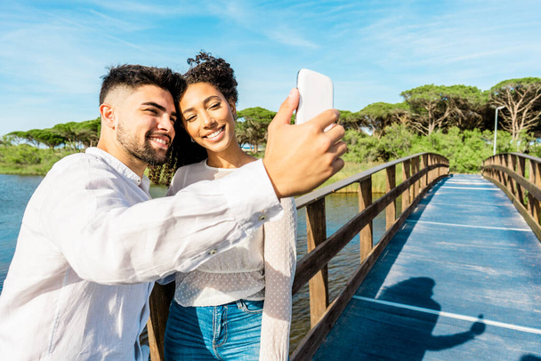 Happy multiracial couple in love staying on a wooden bridge taking a selfie in nature vacations. Handsome guy photographing himself with Hispanic girlfriend using smartphone. New technology habits - Photo, Image