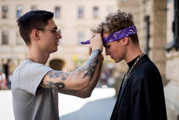 Cute fun curly teenage boy and man wearing stylish 90s fashion clothes with chains around neck. Youth subculture. The man puts a bandana on the guy. Rap artist from new school. - Photo, Image