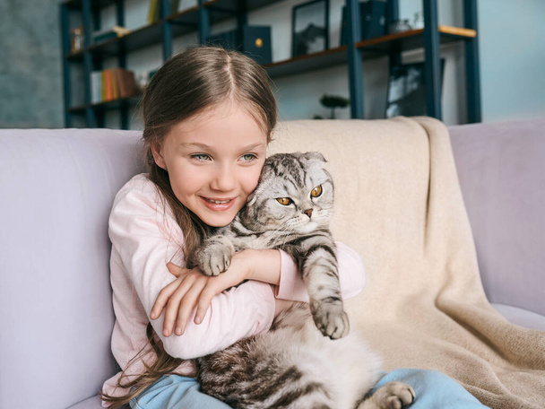 自宅のソファの上にスコットランドの灰色の猫を抱きかかえて、愛らしい、かわいい笑顔の女の子。ペット、友情、家、幸福の概念  - 写真・画像