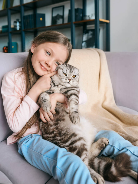 Adorable, mignonne, souriante fille étreignant chat gris écossais sur le canapé à la maison. Animaux, amitié, maison, concept de bonheur  - Photo, image