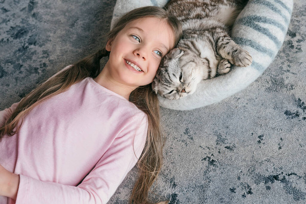 Αξιολάτρευτο, χαριτωμένο, χαμογελαστό κορίτσι ξαπλωμένο με σκωτσέζικη γκρίζα γάτα στο χαλί στο σπίτι. Κατοικίδια ζώα, φιλία, σπίτι, έννοια ευτυχία  - Φωτογραφία, εικόνα